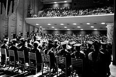 A vintage black and white photograph of IU South Bend graduates seated on stage in a packed auditorium.